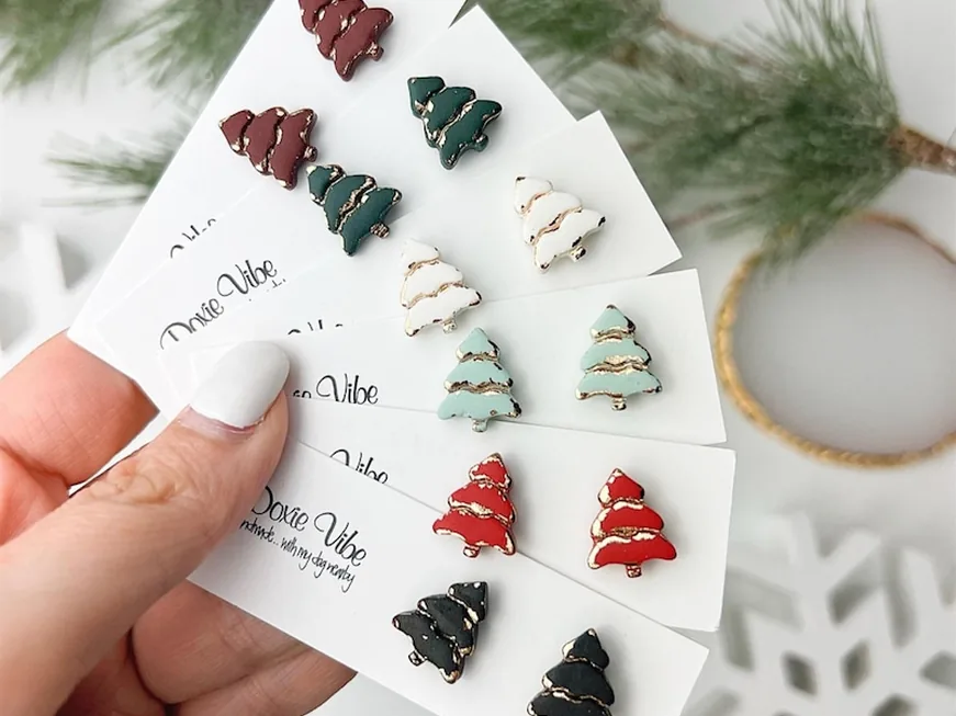 A hand holding cards of tiny Christmas tree stud earrings in red, green and white